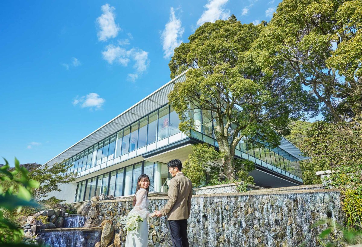 緑と滝に囲まれたSOLA KOBEの外観と、手をつなぐ新郎新婦